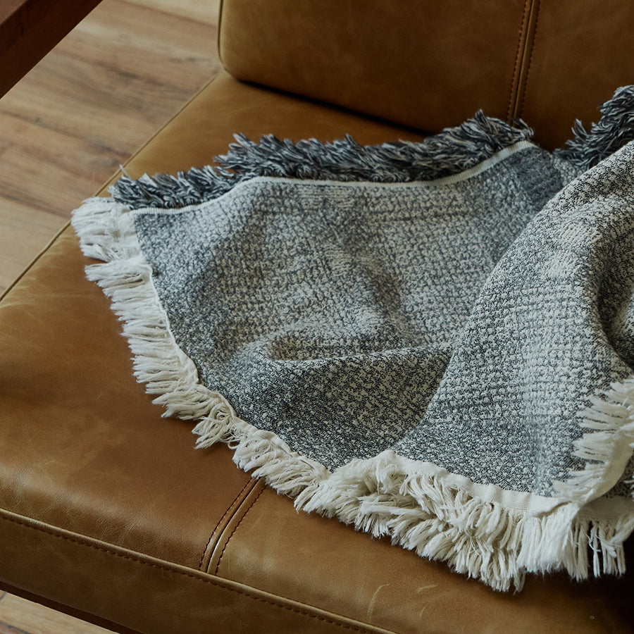 Close-up of the fringe edge on the charcoal black cotton-linen throw resting on a leather chair.