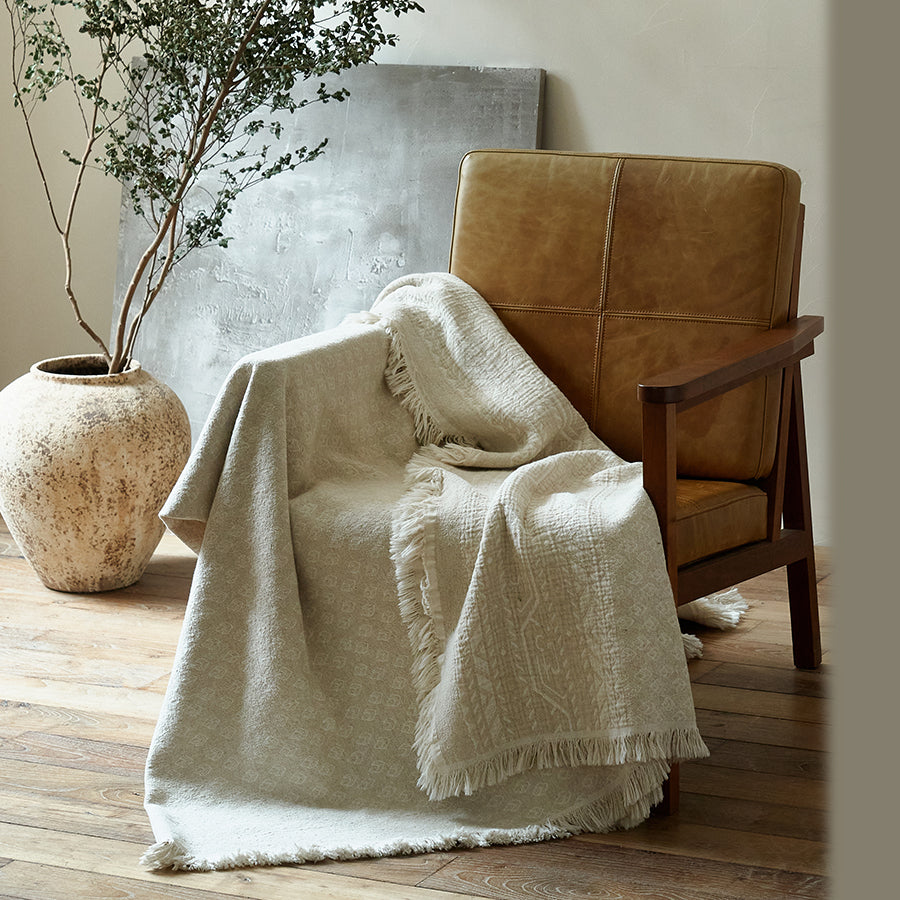Oatmeal beige cotton-linen jacquard blanket draped casually over a brown leather armchair.