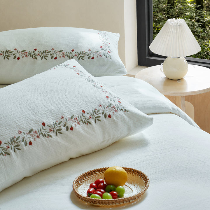 Rosebud Garland embroidered pillow shams displayed on white textured cotton bedding with a bedside lamp.