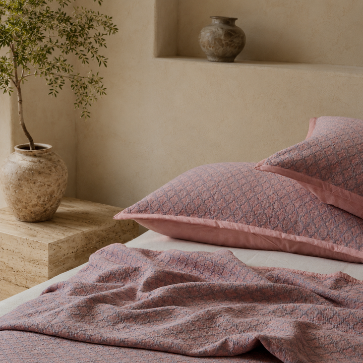 A casual, unmade bed look featuring a dusty rose jacquard pillow and coverlet next to a rustic bedside vase.