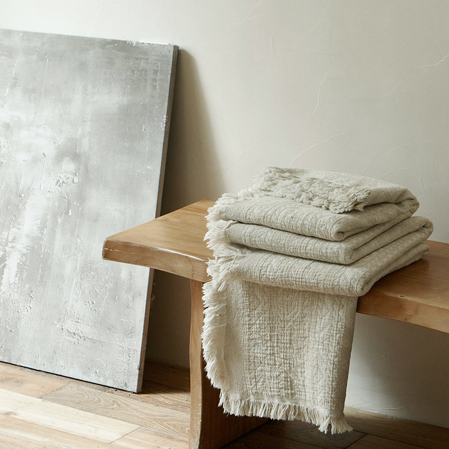 Neatly folded oatmeal beige summer throw blanket resting on a rustic wooden bench.