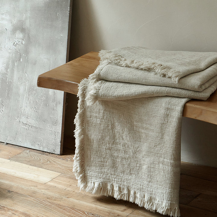 Neatly folded light greige summer throw blanket resting on a minimalist wooden bench.
