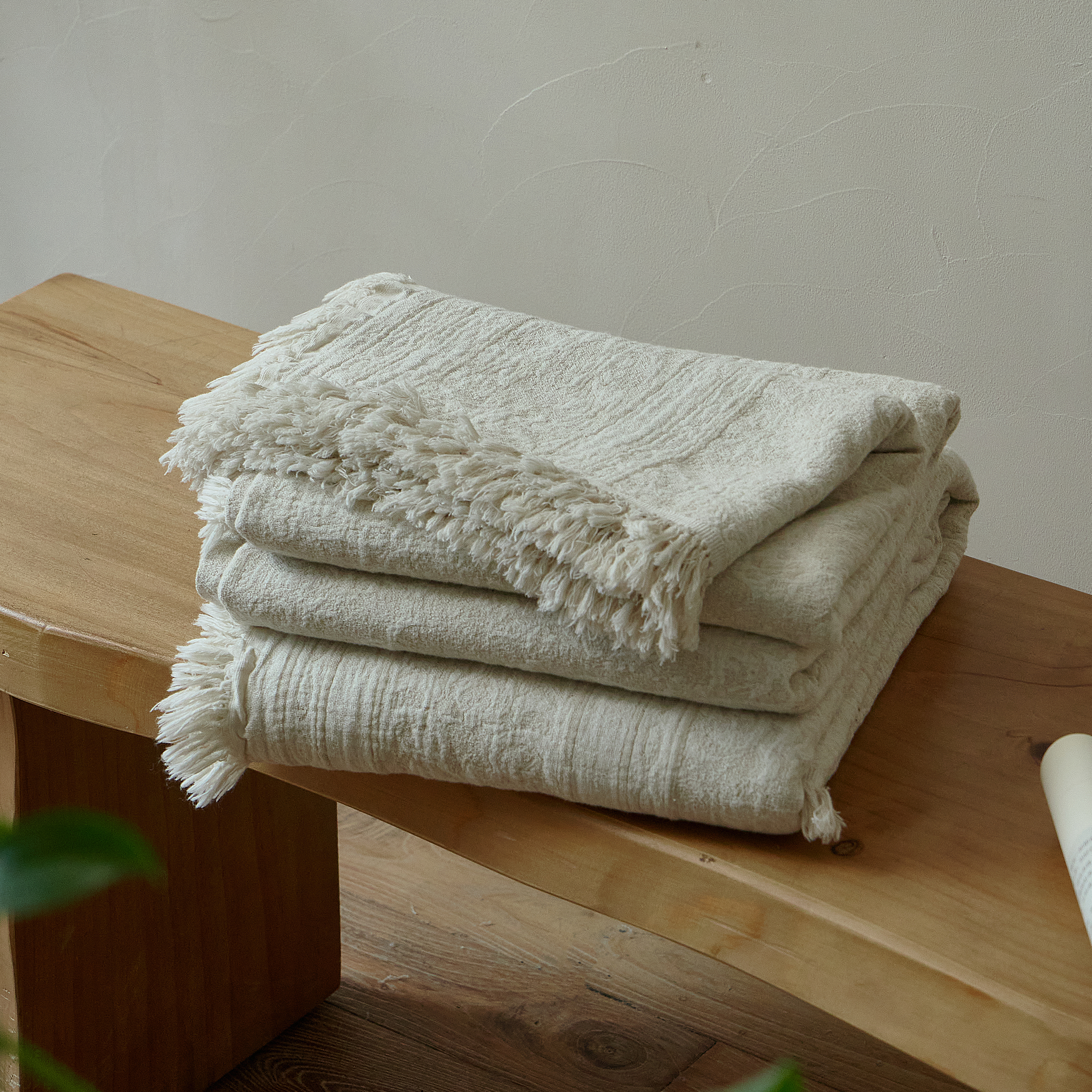 Neatly folded natural ivory cotton-linen blanket with fringed edges resting on a rustic wooden bench.
