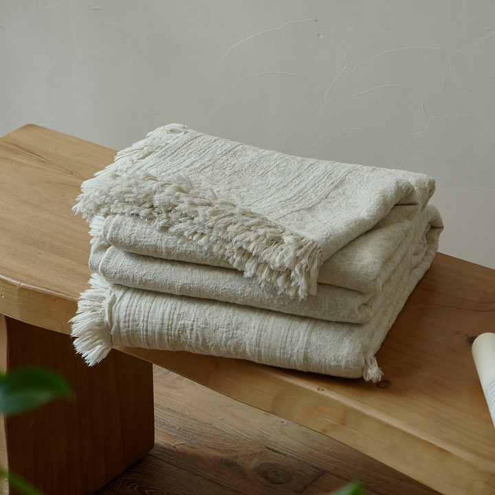 Neatly folded natural ivory cotton-linen blanket with fringed edges resting on a rustic wooden bench.