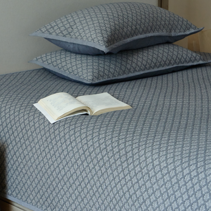 Neatly folded stack of the 3-piece slate blue washed cotton coverlet set with an open book.