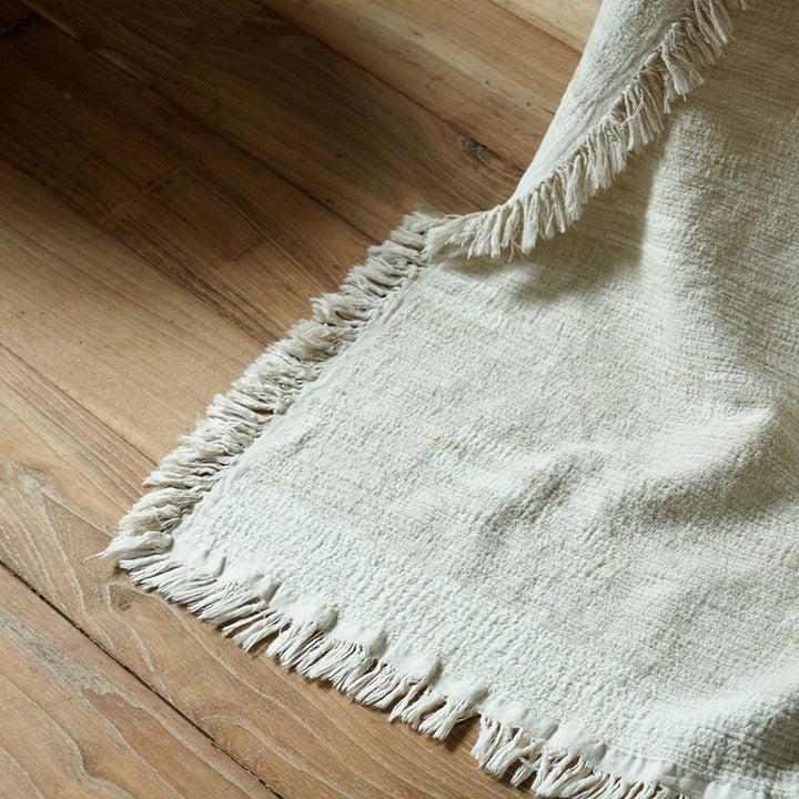 Light greige cotton-linen throw draped over the edge of a bed, trailing beautifully onto a wooden floor.