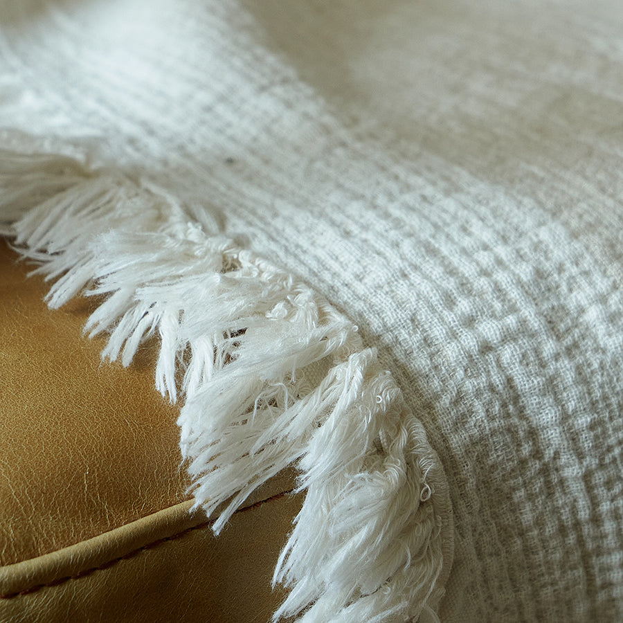 Macro shot of the delicate fringe edge hanging from a light greige linen blend blanket on a leather seat.