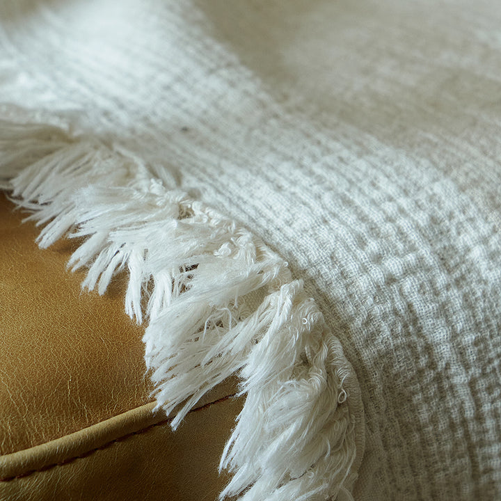 Macro shot of the delicate fringe edge hanging from a light greige linen blend blanket on a leather seat.