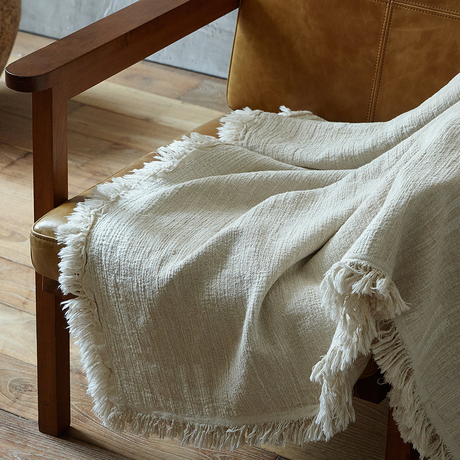 Delicate fringed edge of a light greige textured throw blanket resting gracefully on a wooden chair.