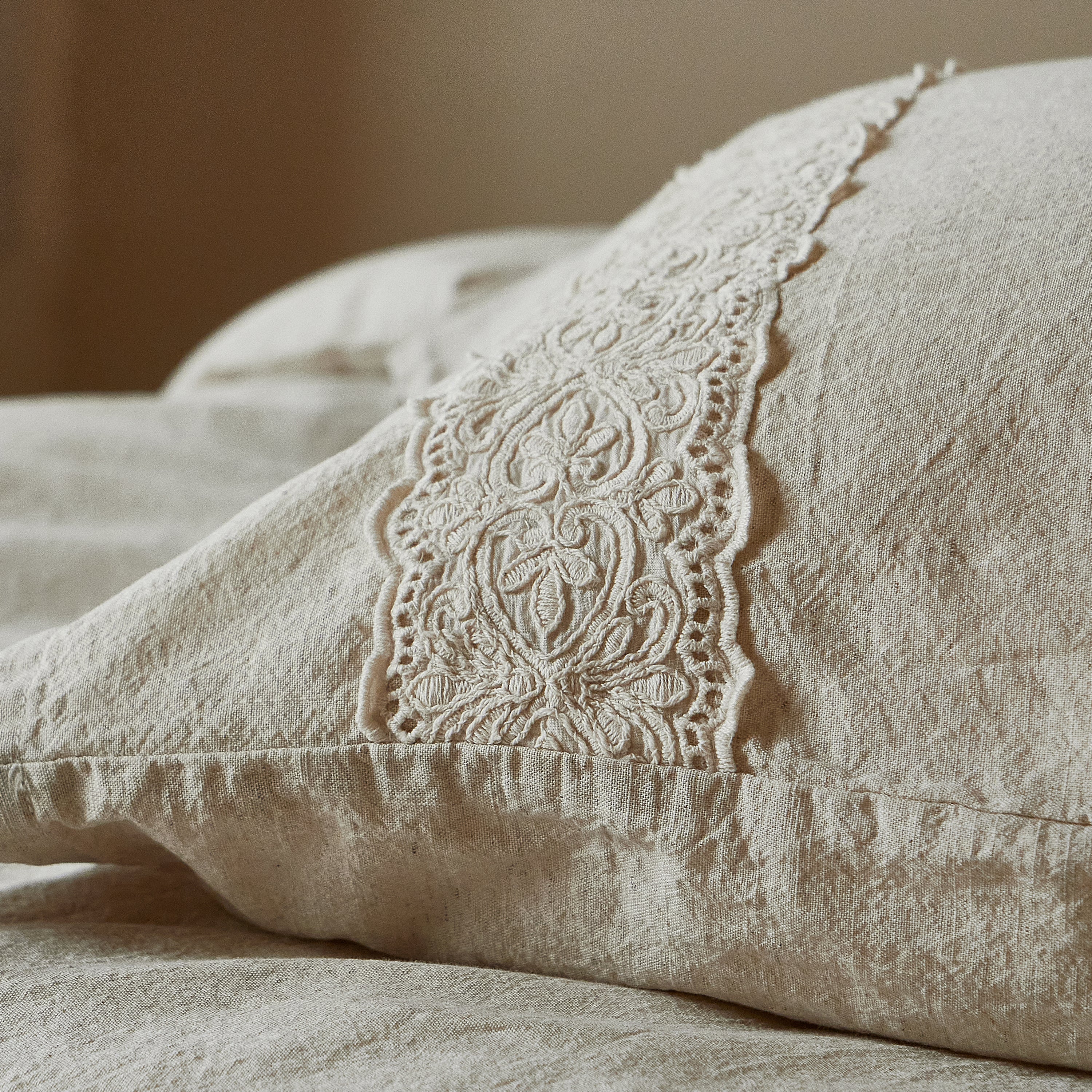 Macro detail of the intricate 3D floral lace embroidery on a natural oatmeal linen pillowcase.