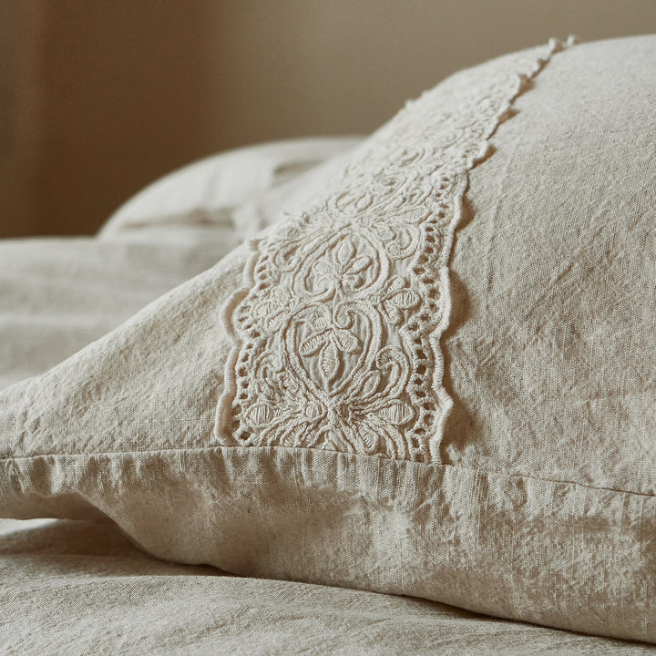 Macro detail of the intricate 3D floral lace embroidery on a natural oatmeal linen pillowcase.