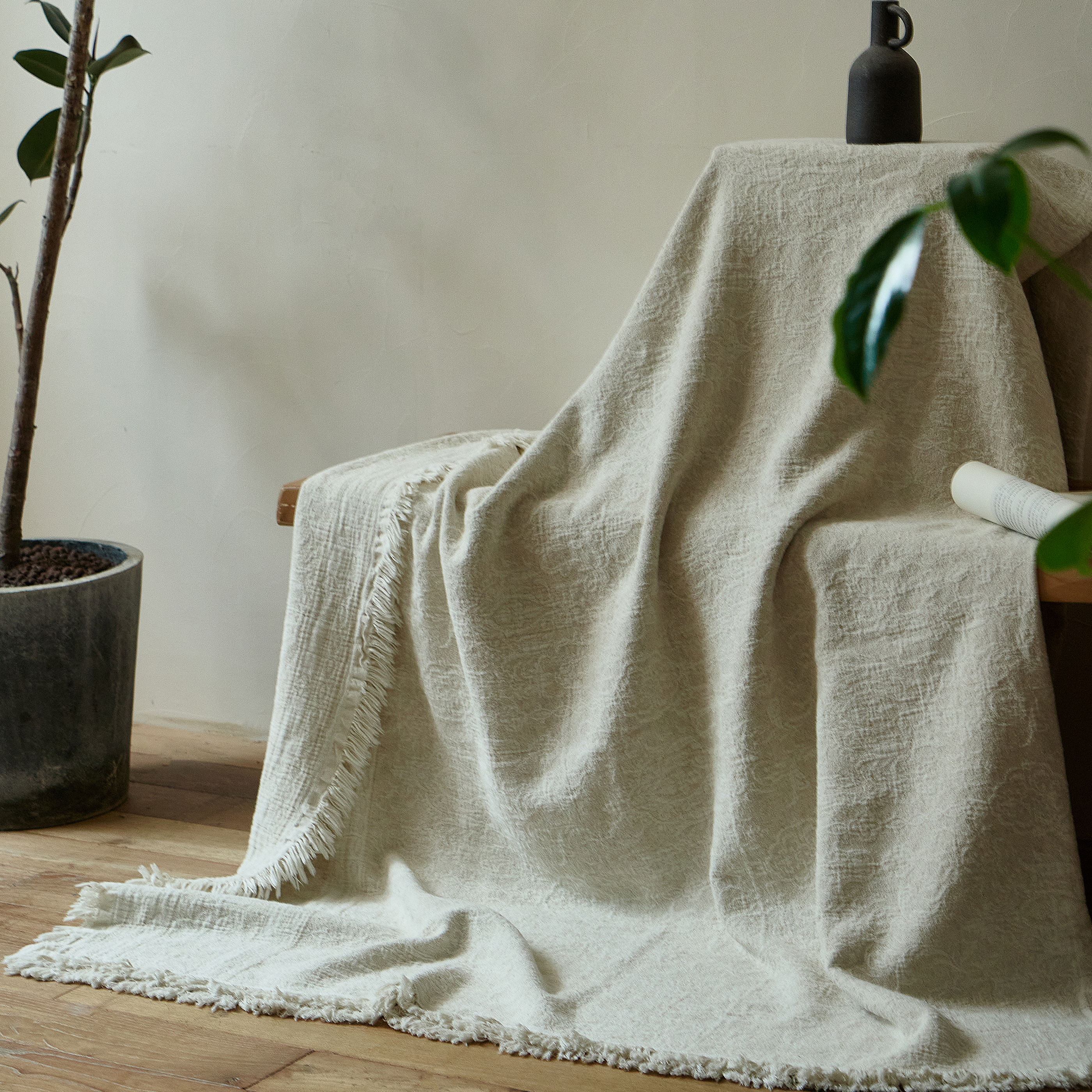 Natural ivory cotton-linen blanket draped gracefully over a chair back, trailing onto a wooden floor next to a plant.