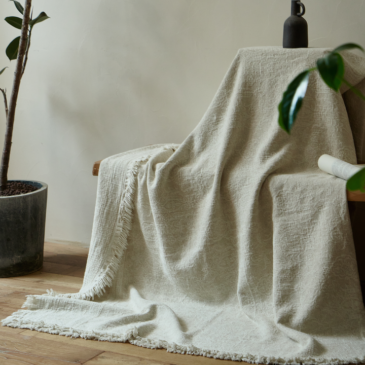 Natural ivory cotton-linen blanket draped gracefully over a chair back, trailing onto a wooden floor next to a plant.