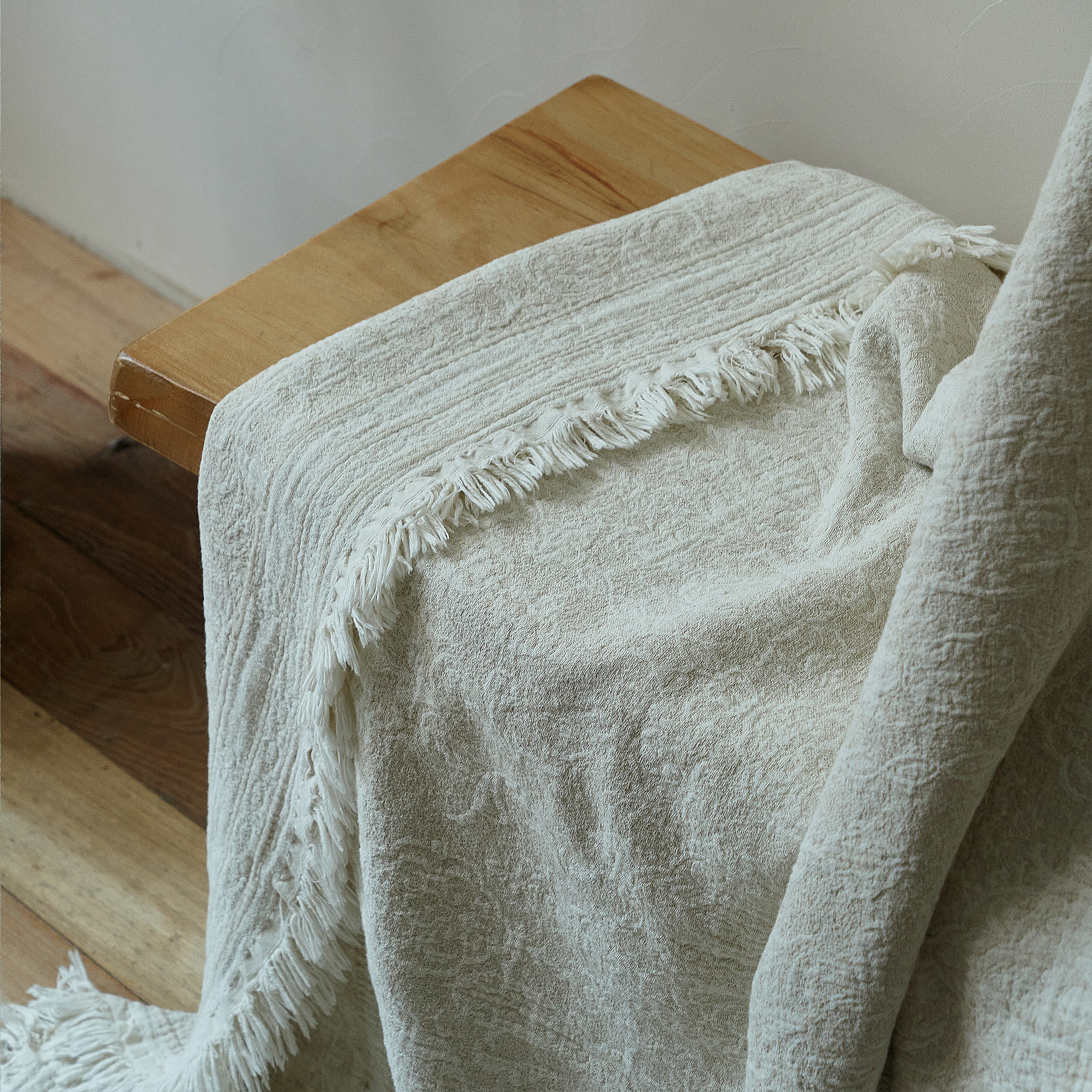 Natural ivory textured throw blanket resting casually on a wooden stool, highlighting the fabric drape.