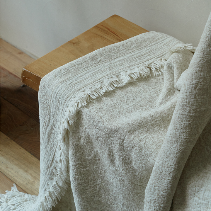 Natural ivory textured throw blanket resting casually on a wooden stool, highlighting the fabric drape.