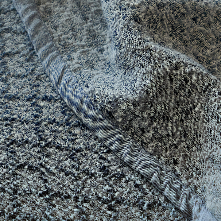 Macro detail of the narrow bound edge and woven texture on the slate blue cotton coverlet.