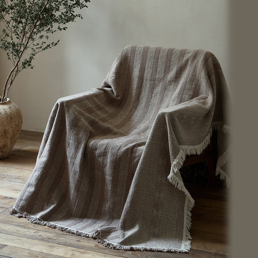 Taupe brown striped cotton-linen blanket draped casually over a chair in a neutral room.