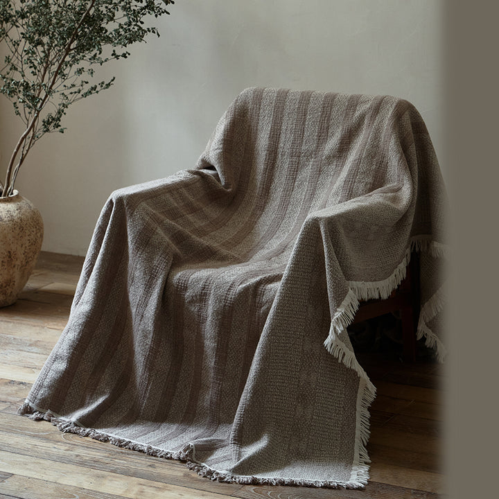 Taupe brown striped cotton-linen blanket draped casually over a chair in a neutral room.