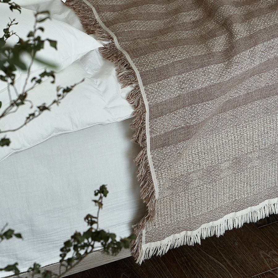 Taupe brown textured throw blanket draped elegantly over white bedding, showing fringe.
