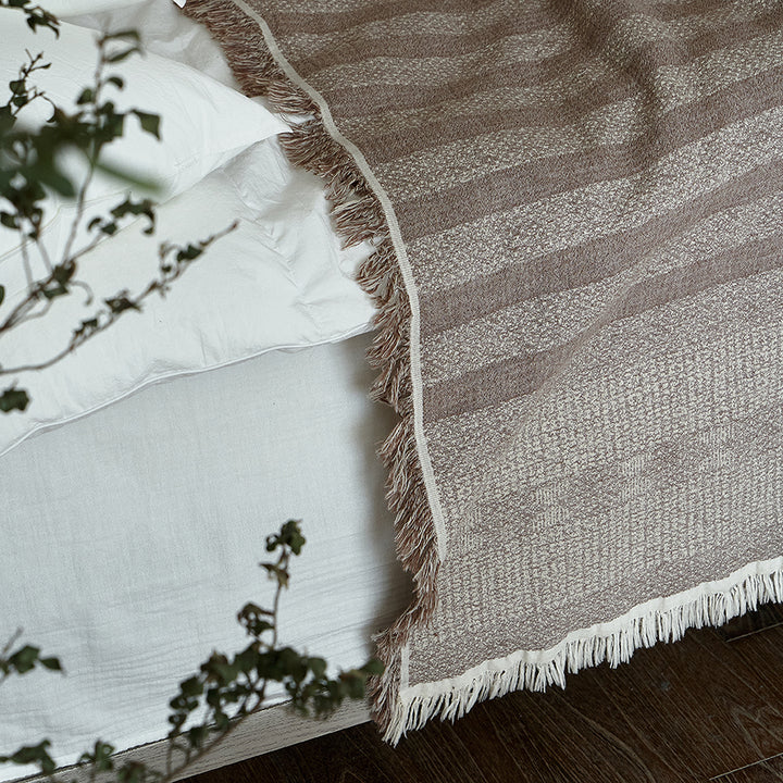 Taupe brown textured throw blanket draped elegantly over white bedding, showing fringe.