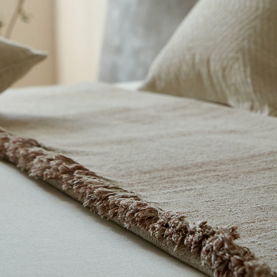 Close-up of the fringed edge of a warm sand cotton-linen throw resting on white bedding.