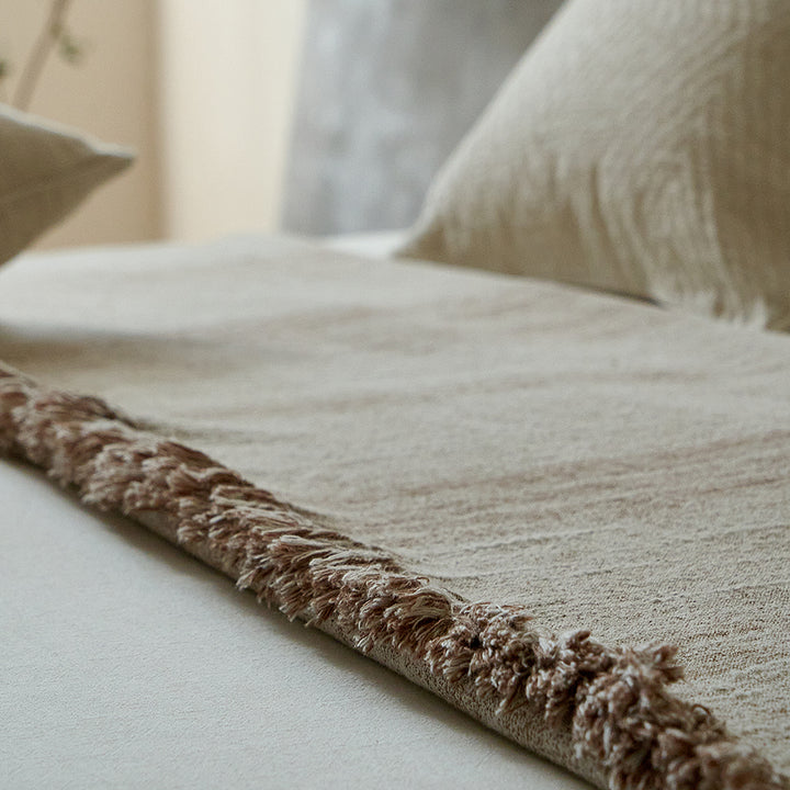 Close-up of the fringed edge of a warm sand cotton-linen throw resting on white bedding.