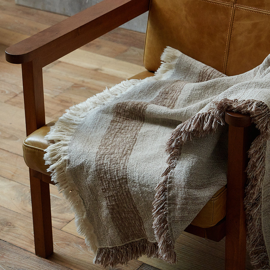 Close-up of the striped border and fringe edge on a warm sand throw resting on a chair arm.